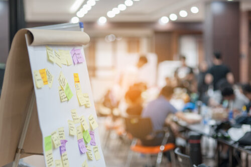 Easel holding poster paper filled with post-it notes, with group of people meeting around a table in the background.