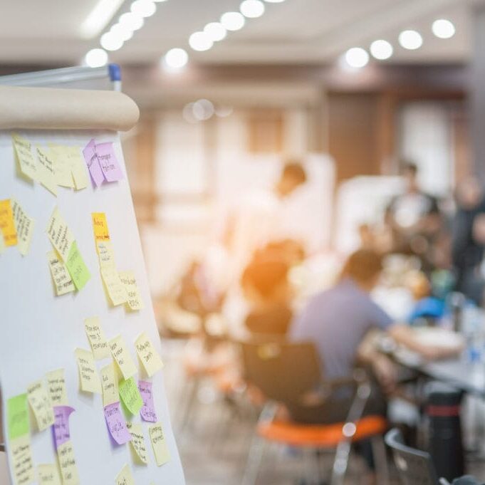 Easel holding poster paper filled with post-it notes, with group of people meeting around a table in the background.