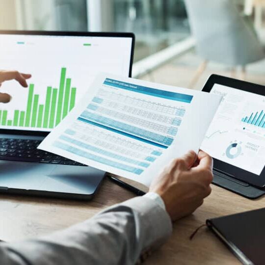 A person compares data on a laptop and a tablet screen with a paper report.